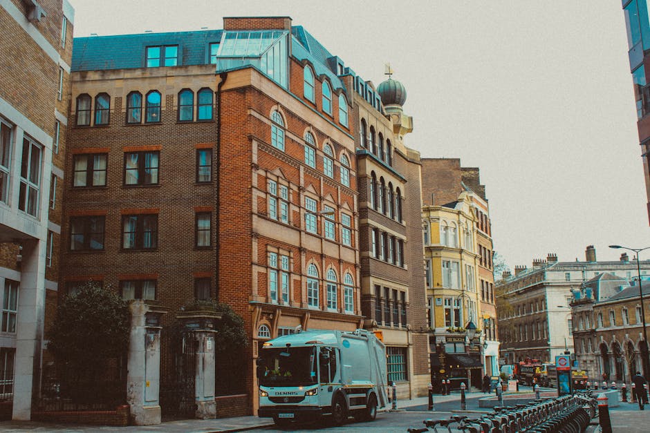 A narrow urban street scene featuring a curved asphalt road with double yellow lines along the edges, leading towards a blue metal bridge structure spanning across the street. On the left side, there