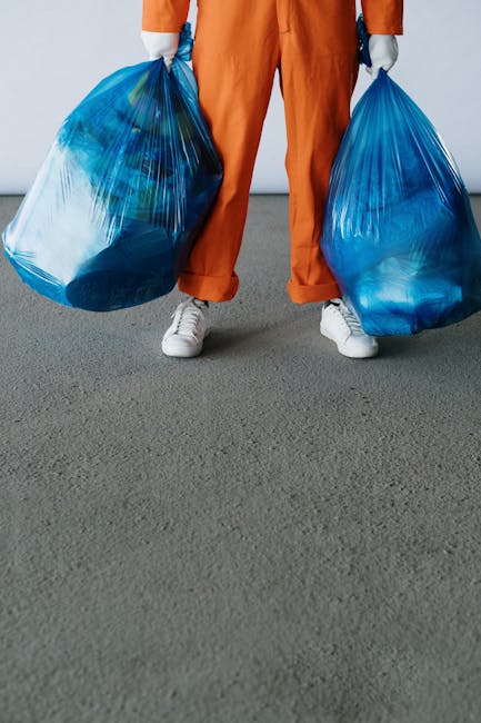 A person wearing an orange protective suit, a face mask with 'End Sars' written on it, and gloves is cleaning debris from underneath a bridge or overpass, using a long-handled brush. The individual is