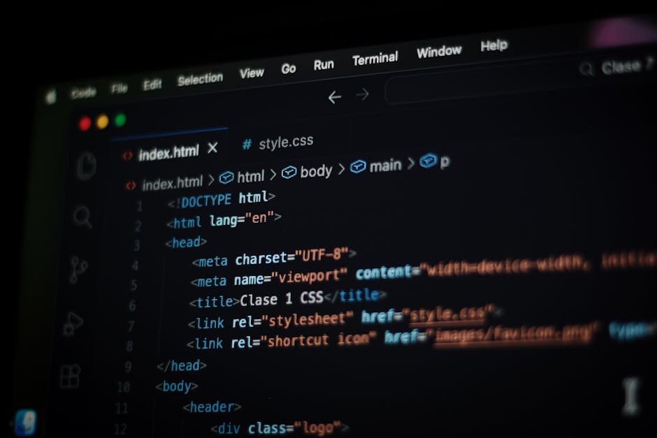 A person with glasses and a beard working at a wooden desk with two large computer monitors displaying lines of code and software interfaces. The workspace includes a wireless keyboard, mouse, and a s